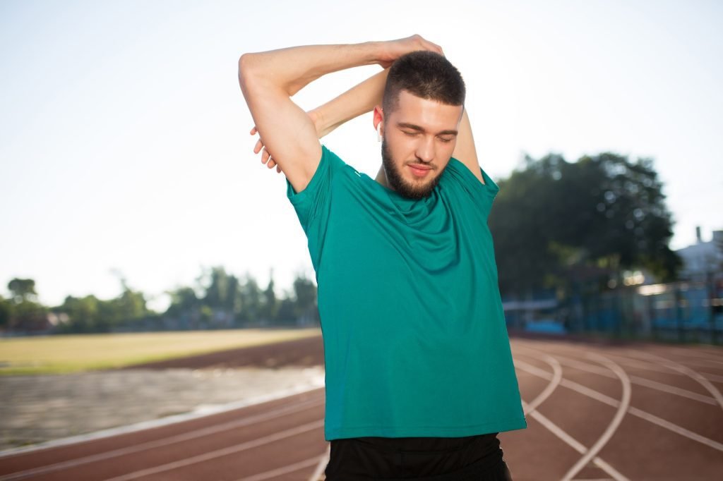 Young man in wireless earphones stretching arms on treadmill of stadium