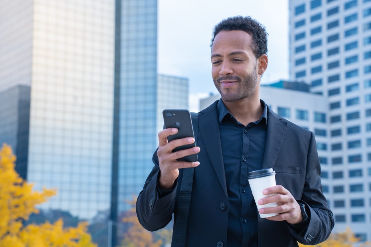 Successful black businessman in suit smiling with smart phone and coffee cup in financial district