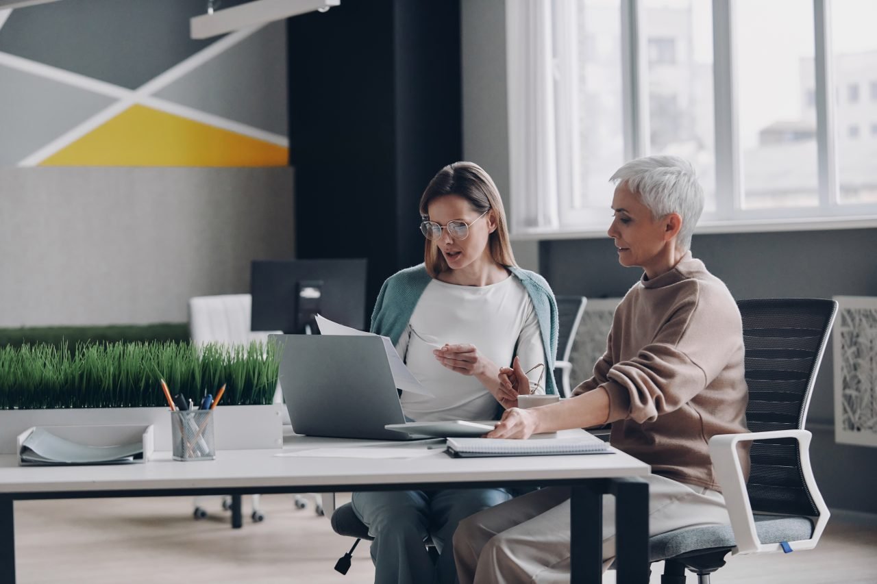 Two confident women using technologies and communicating while working in office together